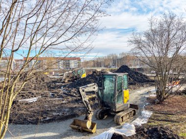 Güneşli bir günde inşaat sahasında çalışan mini kazıcı ve arka planda konut binaları ve diğer makineler.