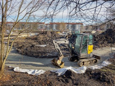 Güneşli bir günde inşaat sahasında çalışan mini kazıcı ve arka planda konut binaları ve diğer makineler.