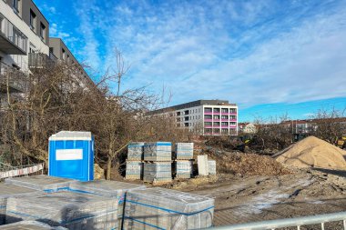 Kum ve çakıl yığınları ve plastik filme sarılmış kaldırım taşı paletleri olan bir inşaat alanı. Ön planda metal bir çit görünür, konut binaları ise görünür.