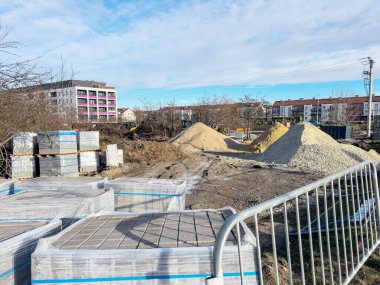 Kum ve çakıl yığınları ve plastik filme sarılmış kaldırım taşı paletleri olan bir inşaat alanı. Ön planda metal bir çit görünür, konut binaları ise görünür.