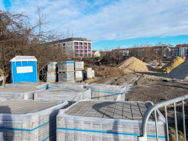 Kum ve çakıl yığınları ve plastik filme sarılmış kaldırım taşı paletleri olan bir inşaat alanı. Ön planda metal bir çit görünür, konut binaları ise görünür.