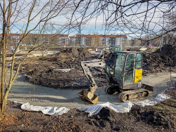 Güneşli bir günde inşaat sahasında çalışan mini kazıcı ve arka planda konut binaları ve diğer makineler.