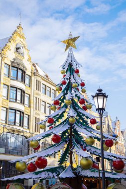 Büyük kırmızı ve altın süslemelerle süslenmiş, karla kaplı dalları taklit eden alışılmadık bir tasarıma sahip Noel ağacı, tarihi şehir binalarına karşı dizilmiş, bayram atmosferi yaratıyor..