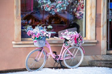 Kış şehrinde bir kafe yakınlarında çiçeklerle süslenmiş pembe bir bisiklet. Estetik çiçek dekoru ve karla romantik bir sokak sahnesi, rahat ve büyüleyici bir şehir atmosferi yaratıyor..