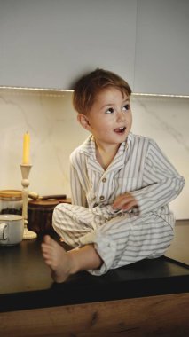 Çizgili pijamalı mutlu çocuk modern bir mutfak tezgahında oturuyor. Rahat ev, çocukluk, sabah, kahvaltı, tarz, masumiyet, duygular. Yüksek kalite fotoğraf