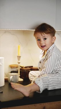 Çizgili pijamalı mutlu çocuk modern bir mutfak tezgahında oturuyor. Rahat ev, çocukluk, sabah, kahvaltı, tarz, masumiyet, duygular. Yüksek kalite fotoğraf