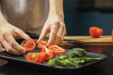 Mutfakta taze bir salata hazırlarken bir tabakta dilimlenmiş domates ve salatalık düzenleyen kişi. Sağlıklı beslenme, taze sebzeler ve evde temiz yemek hazırlığı..