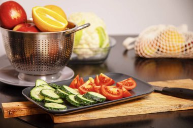 Dilimlenmiş salatalık ve domatesler mutfak masasındaki bir tabakta, arka planda taze meyve ve sebzelerle birlikte. Evde salata hazırlamak için sağlıklı malzemeler.