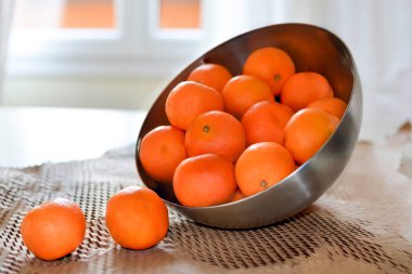 Olgun mandalinalar metal bir kasenin içinde ve masanın üzerinde, pencerenin arka planında yatıyor.