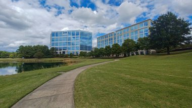 Modern office buildings stand amidst green lawns, trees, and a walking path in Ballantyne Corporate Park under a cloudy sky.