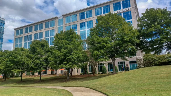 Modern office buildings stand amidst green lawns, trees, and a walking path in Ballantyne Corporate Park under a cloudy sky.