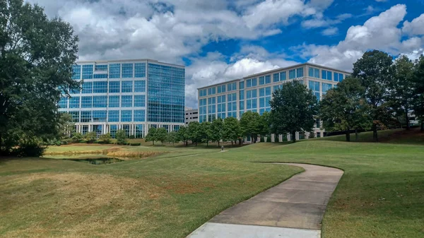 Modern office buildings stand amidst green lawns, trees, and a walking path in Ballantyne Corporate Park under a cloudy sky.