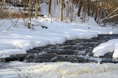 Küçük bir dere karlı bir ormanın içinden akar, karla kaplı kayalar ve kıyılar huzurlu bir kış manzarası yaratır..