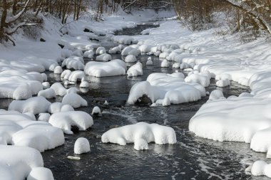 Küçük bir dere karlı bir ormanın içinden akar, karla kaplı kayalar ve kıyılar huzurlu bir kış manzarası yaratır..