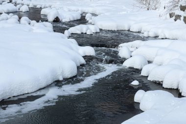 Küçük bir dere karlı bir ormanın içinden akar, karla kaplı kayalar ve kıyılar huzurlu bir kış manzarası yaratır..