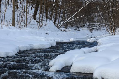 Küçük bir dere karlı bir ormanın içinden akar, karla kaplı kayalar ve kıyılar huzurlu bir kış manzarası yaratır..
