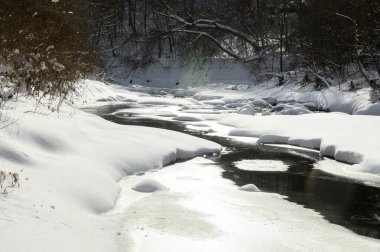 Küçük bir dere karlı bir ormanın içinden akar, karla kaplı kayalar ve kıyılar huzurlu bir kış manzarası yaratır..