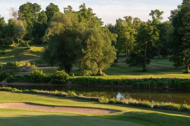 Manzaralı bir golf sahasında güneşli bir günde yeşil bir sığınak ve ağaçlar bulunur..