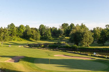 Manzaralı bir golf sahasında güneşli bir günde yeşil bir sığınak ve ağaçlar bulunur..