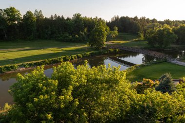 Gün batımında güzel yeşil bir parkta sakin bir nehri geçen bir yaya köprüsü..