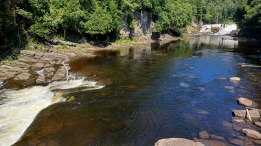 Sakin nehir açık mavi gökyüzünü yansıtır ve güneşli bir öğleden sonra yemyeşil orman ve kayalık kayalıklarla çevrilidir..