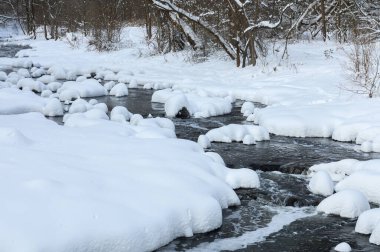 Küçük bir dere karlı bir ormanın içinden akar, karla kaplı kayalar ve kıyılar huzurlu bir kış manzarası yaratır..