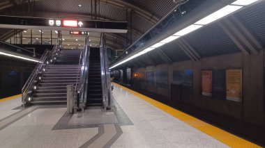 Toronto, ON, Kanada - 31 Ekim 2025: Toronto Transit Komisyonu 'nun (TTC) metro istasyonunun boş bir metro platformu, üst kata çıkan uzun bir yürüyen merdiven ve merdiven ve mesafeye giden bir tren ile birlikte..