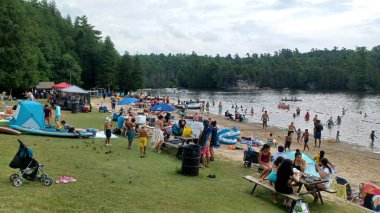 Rockwood, ON, Kanada - 19 Ağustos 2025: Büyük bir kalabalık bulutlu bir yaz gününde çimlerin üzerinde rahatlar ve gölde yüzer..