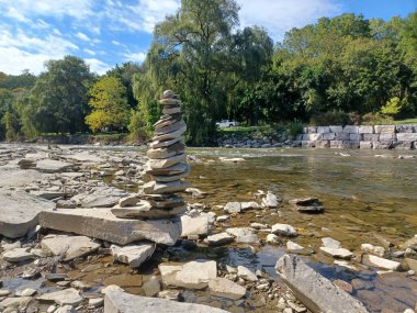 Uzun, dengeli bir taş cairn, akan bir nehrin kayalık kıyısında duruyor. Parlak mavi gökyüzünün altında yeşil ağaçlar var..