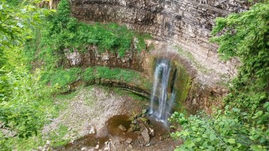 Küçük bir Taws şelalesi, doğal park ortamındaki yemyeşil ormanlarla çevrili katmanlı kayalık yamaçların üzerinde çağlıyor..