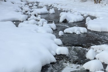 Küçük bir dere karlı bir ormanın içinden akar, karla kaplı kayalar ve kıyılar huzurlu bir kış manzarası yaratır..
