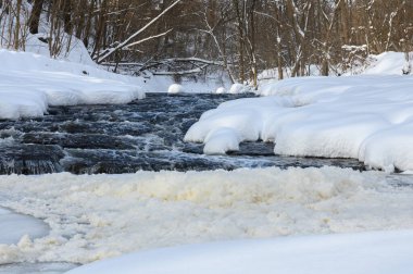 Küçük bir dere karlı bir ormanın içinden akar, karla kaplı kayalar ve kıyılar huzurlu bir kış manzarası yaratır..