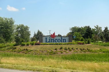 Lincoln, ON, Kanada - 18 Eylül 2025: Mavi gökyüzü altında yemyeşil ağaçların ve yemyeşil ağaçların arasında kırmızı bir akçaağaç yaprağı amblemi bulunan önemli bir giriş işareti.