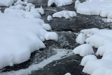 Küçük bir dere karlı bir ormanın içinden akar, karla kaplı kayalar ve kıyılar huzurlu bir kış manzarası yaratır..