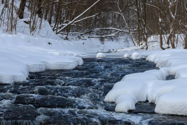 Küçük bir dere karlı bir ormanın içinden akar, karla kaplı kayalar ve kıyılar huzurlu bir kış manzarası yaratır..