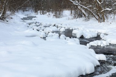Küçük bir dere karlı bir ormanın içinden akar, karla kaplı kayalar ve kıyılar huzurlu bir kış manzarası yaratır..
