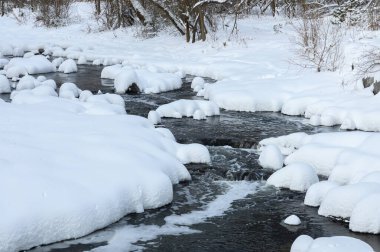 Küçük bir dere karlı bir ormanın içinden akar, karla kaplı kayalar ve kıyılar huzurlu bir kış manzarası yaratır..