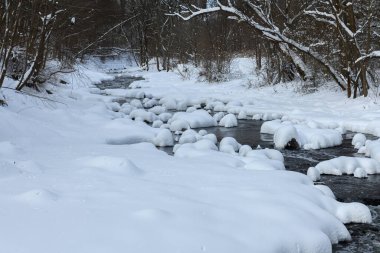 Küçük bir dere karlı bir ormanın içinden akar, karla kaplı kayalar ve kıyılar huzurlu bir kış manzarası yaratır..