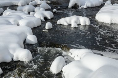 Küçük bir dere karlı bir ormanın içinden akar, karla kaplı kayalar ve kıyılar huzurlu bir kış manzarası yaratır..