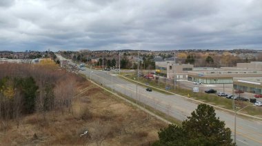 Yüksek bir manzara banliyö manzarasını, çıplak kış ağaçlarını, trafiğin bitişik olduğu bir yolu ve parçalı bulutlu bir gökyüzünün altındaki uzak şehir binalarını gösterir..