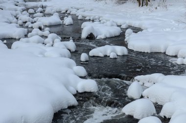 Küçük bir dere karlı bir ormanın içinden akar, karla kaplı kayalar ve kıyılar huzurlu bir kış manzarası yaratır..