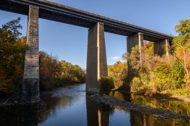 Uzun sütunları olan beton köprü yapısı Etienne Brule Parkı 'ndaki açık mavi gökyüzü altında canlı sonbahar yapraklarıyla çevrili, sakin Humber Nehri üzerinde bir demiryolu hattı taşıyor.