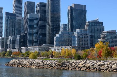 Etobicoke, Toronto, Kanada 'da panoramik göl silüeti oluşturan modern yerleşim binaları, park boyunca canlı sonbahar yaprakları ve açık mavi gökyüzü altında kayalık kıyı şeridi oluşturur.