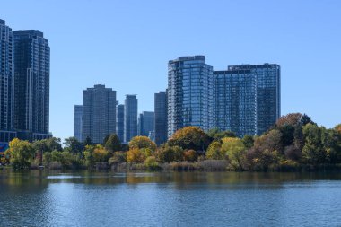 Etobicoke, Toronto 'daki modern yerleşim yerleri, berrak mavi gökyüzünün altında sakin bir göl etrafında uzanan canlı sonbahar ağaçlarının arkasında dikiliyor, kentsel doğal güzellikleri gösteriyor.