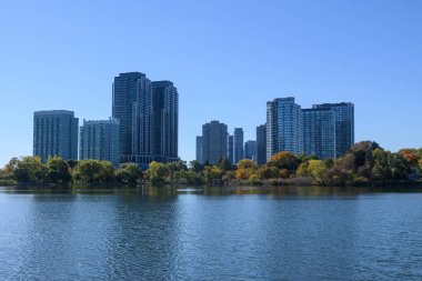 Etobicoke Toronto silueti, ağaçlarla kaplı bir rıhtımın üzerinde yükselen modern yüksek binaları ve derin mavi gökyüzüne ve yansıtıcı göle karşı canlı sonbahar yapraklarını gösteriyor.