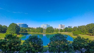 Charlotte, NC, ABD - 28 Nisan 2025: Berrak bir gökyüzü altında modern ofis ve yerleşim binalarıyla çevrili yemyeşil ağaçlar ve küçük bir gölet.