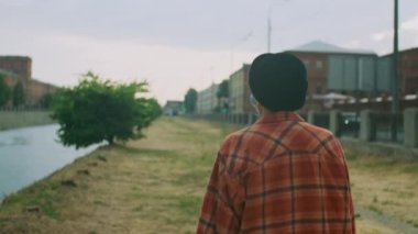 Koruyucu tıbbi maske takan koyu renk saçlı güzel bir kızın arkadan çekimini takip et. Güzel bir şapka. Kıyı şeridinde gezen şık elbiseli bir kadın. Bulanık şehir arkaplanı