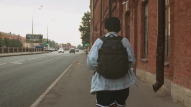 Koyu renk saçlı, güzel bir kızın arka plan fotoğrafını takip et. Şapka ve sırt çantası takan, şehirde e-scooter kullanan. Bulanık kasaba geçmişi. Kırmızı tuğla duvar. Yoldan geçen arabaların yanından