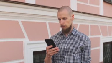 Kel sakallı adam akıllı telefon kullanarak sosyal medyada sörf yapıyor, haberleri kontrol ediyor, oyun oynuyor, iş görüşmesi yapıyor ya da güneşli bir günde sokakta mesaj yazıyor. bina duvarı arkaplanı