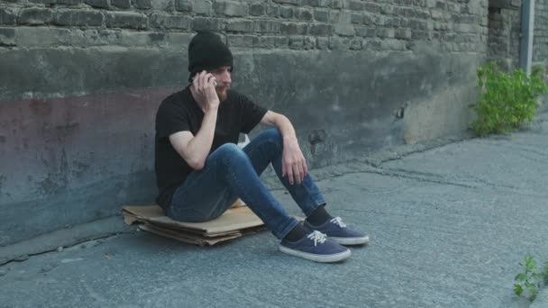 ホームレスの男が通りの段ボール箱の上に座って 彼の電話を壊します 貧困と惨めさに陥り解雇され解雇され解雇された 社会問題失業概念 — ストック動画